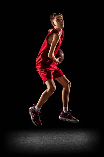 Full längd porträtt av pojke, tonåring, professionell basketspelare scoring mål i hopp, kasta bollen i korg isolerad över svart bakgrund — Stockfoto