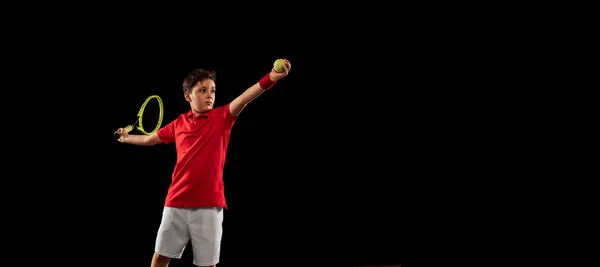 Full-length portrait of young boy, tennis player in motion, training, serving ball isolated over black background. Concentration — Stock Photo, Image