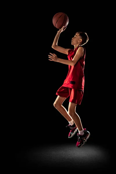 Ganzkörperporträt eines sportlichen Teenagers, Basketballspielers in roter Uniform, der Ball isoliert über schwarzem Hintergrund in den Korb wirft — Stockfoto