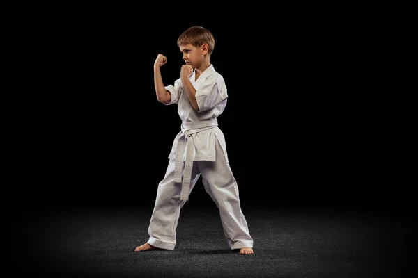 Portret van kleine jongen, karate sportman in witte kimono staan met vuisten omhoog geïsoleerd over zwarte achtergrond — Stockfoto