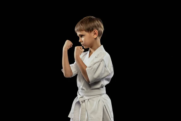 Портрет маленького хлопчика в білому кімоно тренування бойових мистецтв, що стоїть з кулаками вгору ізольовано на чорному тлі — стокове фото