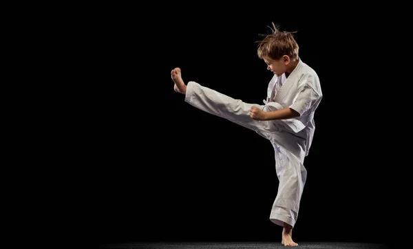 Retrato completo de niño pequeño, karateka, entrenamiento de arte marcial aislado sobre fondo negro. Posición de patada lateral — Foto de Stock