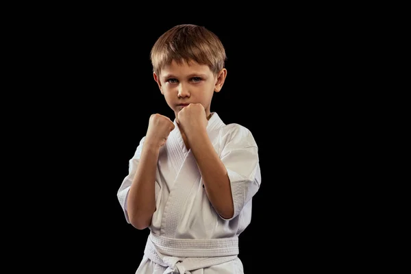 Retrato de niño pequeño en kimono blanco entrenando karate aislado sobre fondo negro. Puños arriba —  Fotos de Stock