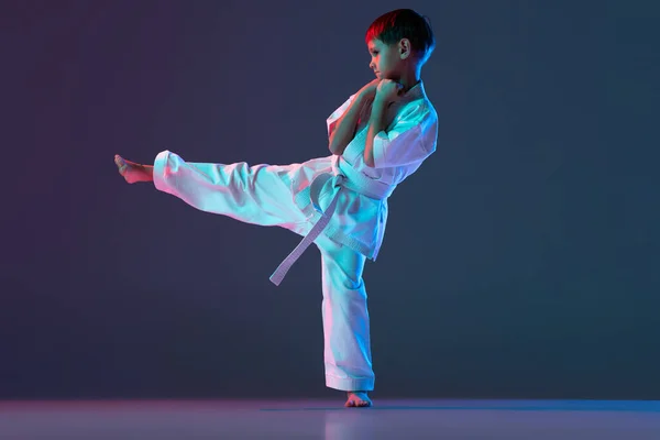 Sportos gyermek teljes hosszúságú portréja, edzés karate ütés elszigetelt gradiens kék háttér a neon — Stock Fotó