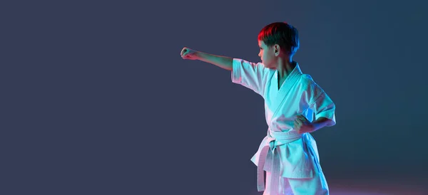 Przycięty portret chłopca, karateka trening przodu pozycja odizolowana nad gradientowym niebieskim tle fioletowym w neonie. Ulotka — Zdjęcie stockowe