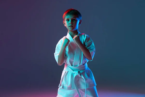 Portret van kleine jongen in witte kimono training karate geïsoleerd over gradiënt blauwe achtergrond in neon. Vuisten omhoog — Stockfoto