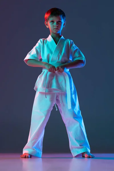 Retrato completo de menino, karateka em quimono branco posando isolado sobre gradiente fundo azul em luzes de néon — Fotografia de Stock