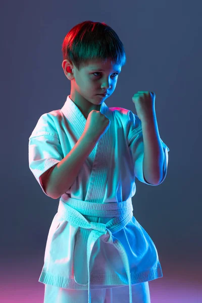 Retrato cortado de criança, treinamento esportivo concentrado, praticando karatê isolado sobre fundo azul gradiente em néon — Fotografia de Stock