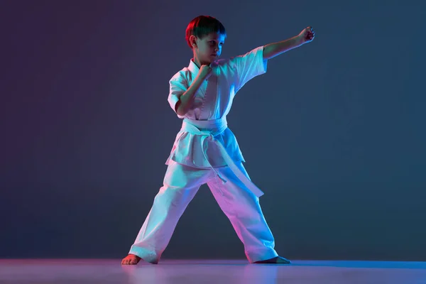 Πλήρες μήκος πορτρέτο του παιδιού, αθλητή κατάρτισης καράτε θέτει απομονωμένη πάνω κλίση μπλε μωβ φόντο σε νέον — Φωτογραφία Αρχείου