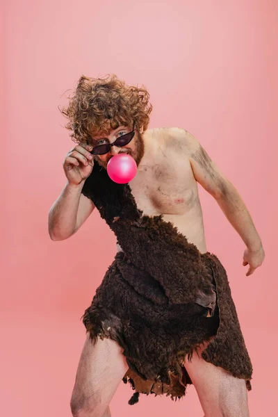 Portrait d'homme artistique de caractère néandertalien portant une peau d'animal, des lunettes de soleil élégantes et mangeant de la gomme à bulles isolée sur fond rose — Photo