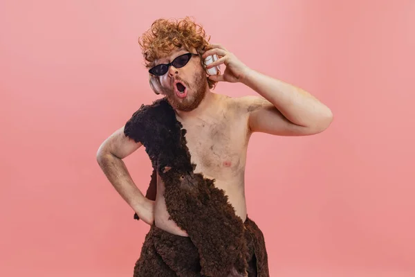 Portrait recadré de l'homme en caractère néandertalien portant des lunettes de soleil élégantes et écoutant de la musique dans des écouteurs isolés sur fond rose — Photo