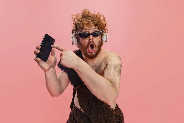 Portrait de l'homme dans le caractère de néandertal dans les écouteurs pointant vers le téléphone avec une expression choquée isolé sur fond rose — Photo