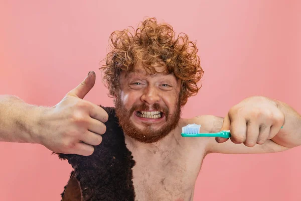 Portrait recadré d'homme drôle dans le caractère de dents brossant néandertal avec du dentifrice isolé sur fond rose. Montrant comme geste — Photo