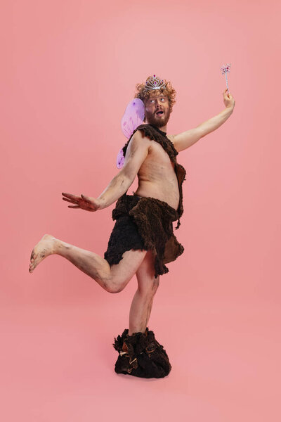 Portrait of man in character of neanderthal wearing fairy wings and crown posing like little wizzard isolated over pink background. CVute