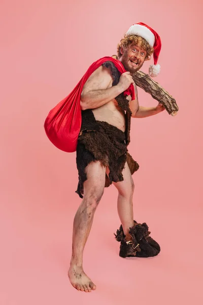 Retrato completo de homem alegre, neandertal usando chapéu de Natal, segurando log e saco vermelho com presentes isolados sobre fundo rosa — Fotografia de Stock