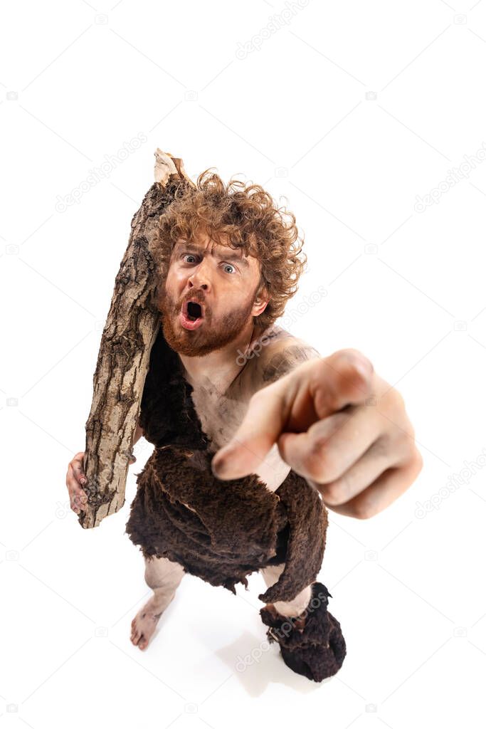 Full-length portrait of man in character of neanderthal, primitive person wonderingly pointing at camera isolated over white background