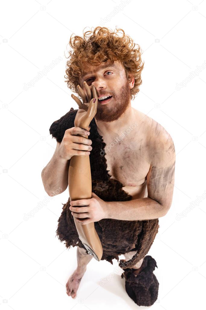Full-length portrait of man in character of neanderthal holding fake human hand and putting finger into nose isolated over white background