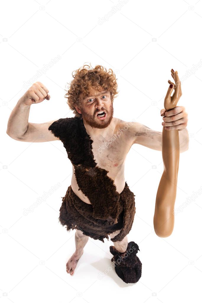 Full-length portrait of man in character of neanderthal holding fake human hand and showing muscle strenght isolated over white background