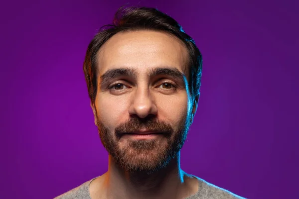 Крупным планом портрет человека внимательно смотрит в камеру, позируя изолированным на фиолетовом фоне в неоновом свете — стоковое фото