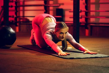 Genç sportif kız eğitiminin tam boy portresi, sırt üstü uzanış, beden eğitimi arka planında izole edilmiş halı üzerinde vücut germe.