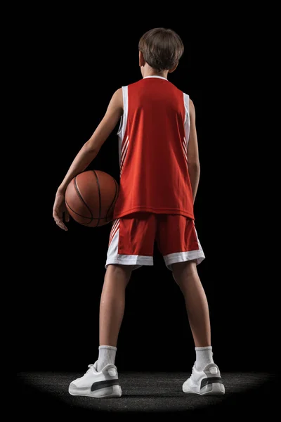 Retrato completo de menino adolescente, jogador de basquete em uniforme vermelho posando com bola isolada sobre fundo preto. Visão traseira — Fotografia de Stock