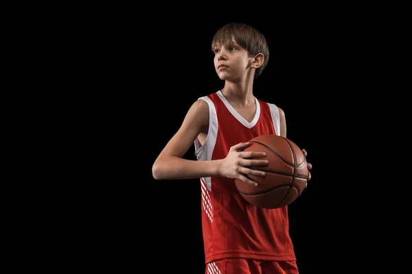 Cropped portrait of young boy, basketball player in red uniform standing, posing with ball isolated over black background — Stock Photo, Image