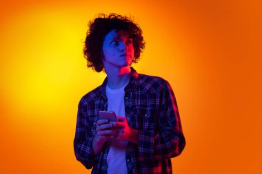 Genç kıvırcık adamın portresi, kareli gömlekli öğrenci düşünceli görünüyor ve turuncu arka planda izole edilmiş bir şekilde telefon mesajı yazıyor.