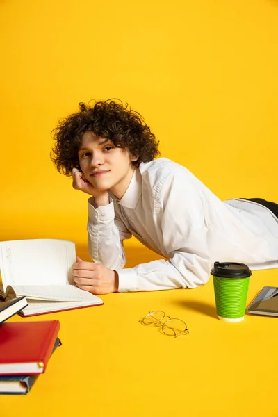 Portré fiatal fiúról, egy diákról, aki a földön fekszik, könyvet olvas, házi feladatot csinál, sárga háttérrel elszigetelve. Nézem a kamerát, pózolok — Stock Fotó