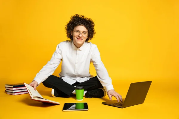 Retrato de jovem sorridente homem, estudante olhando para a câmera, posando em torno de laptop e livros isolados sobre fundo estúdio amarelo — Fotografia de Stock