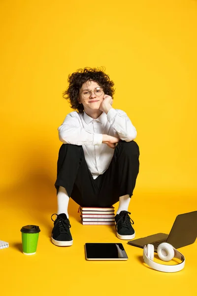 Retrato de jovem sorridente, estudante sentado em livros e olhando para a câmera, posando isolado sobre fundo de estúdio amarelo — Fotografia de Stock
