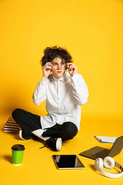 Retrato de jovem, estudante de óculos sentado no chão em torno de livros, laptop e tablet isolado sobre fundo estúdio amarelo — Fotografia de Stock