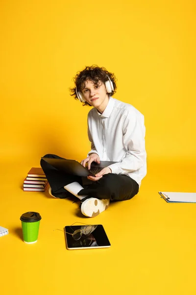 Retrato de jovem homem encaracolado, estudante ouvindo aulas on-line em heaphones no laptop isolado sobre fundo estúdio amarelo — Fotografia de Stock