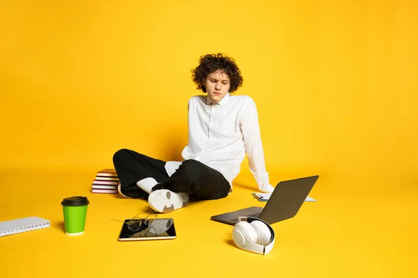 Um jovem, estudante sentado no chão, olhando para o laptop, tendo aulas on-line isolado sobre fundo estúdio amarelo — Fotografia de Stock