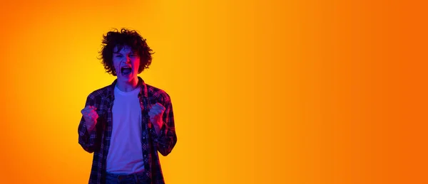 Porträt eines jungen lockigen Mannes, Student im karierten Hemd, der Emotionen der Aufregung isoliert vor orangefarbenem Studiohintergrund in blauem Neon zeigt — Stockfoto
