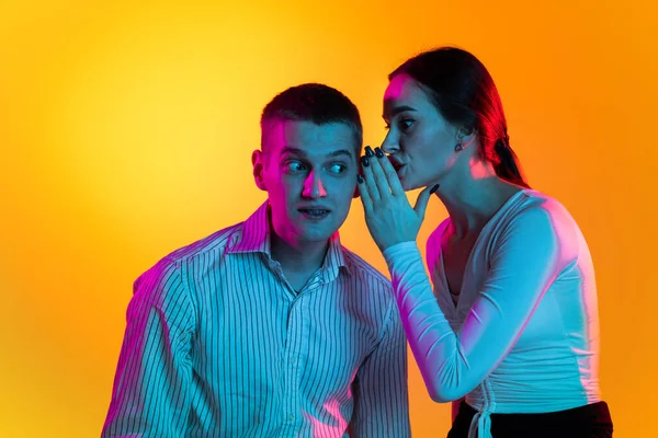 Portret van twee jonge mensen, vrouw fluisterend op het oor van een man, pratend over nieuws geïsoleerd over oranje achtergrond in neonlicht — Stockfoto