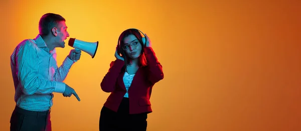 Portrait of young man shouting in megaphone at woman in headphones isolated over orange studio background in neon lights — ストック写真