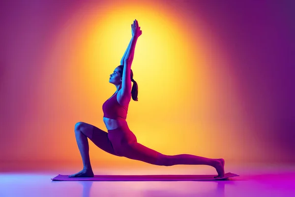 Portrét mladé fit dívka v červené teplákové soupravě dělá jóga strečink cvičení izolované přes gradient růžové a žluté pozadí v neonu — Stock fotografie