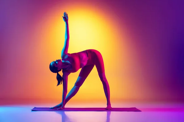 Portret van jong sportief meisje in rode trainingspak, stretching oefeningen geïsoleerd over gradiënt roze en gele achtergrond in neon — Stockfoto