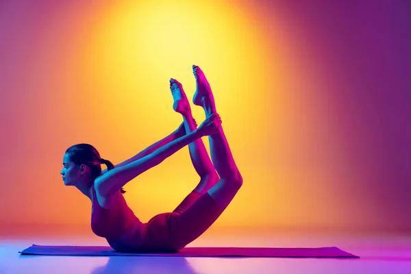 Portrait de jeune fille active faisant des exercices de yoga, tenant les jambes avec les mains isolées sur dégradé fond rose et jaune au néon. — Photo