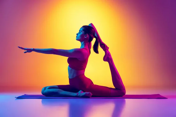 Portrét mladé sportovní ženy v černé teplákové soupravě dělá jógu a strečink cvičení izolované přes gradient růžové a žluté pozadí v neonu — Stock fotografie