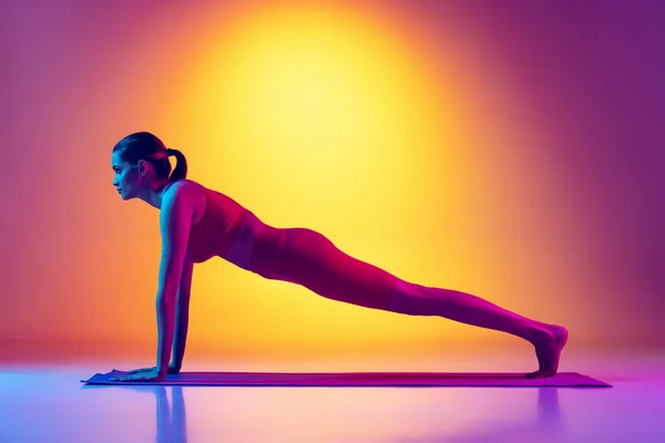 Une jeune fille en survêtement rouge faisant des exercices d'étirement de yoga, debout dans une planche isolée sur fond rose et jaune dégradé au néon — Photo