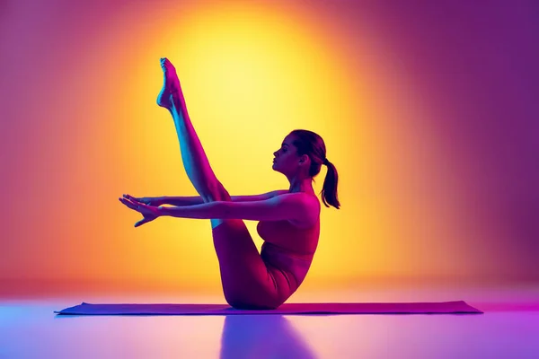 Portrét mladé sportovní dívky trénink, sezení, stoupající noha nahoru s rovnými pažemi izolované přes gradient růžové a žluté pozadí v neonu. — Stock fotografie