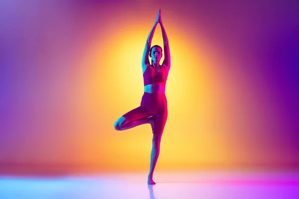 Портрет молодой женщины, делающей упражнения на растяжку и йогу изолированные на градиентном розовом и желтом фоне в неоновом — стоковое фото
