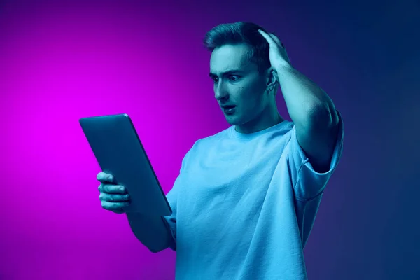 Portrait of young man reading news on tablet with shocked expression isolated over gradient pink purple background in blue neon — Stock Photo, Image