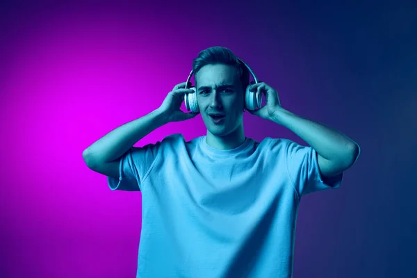 Porträt eines jungen emotionalen Mannes, der Musik mit Kopfhörern hört, isoliert über einem Gradienten in rosa und lila Hintergrund in Neon — Stockfoto