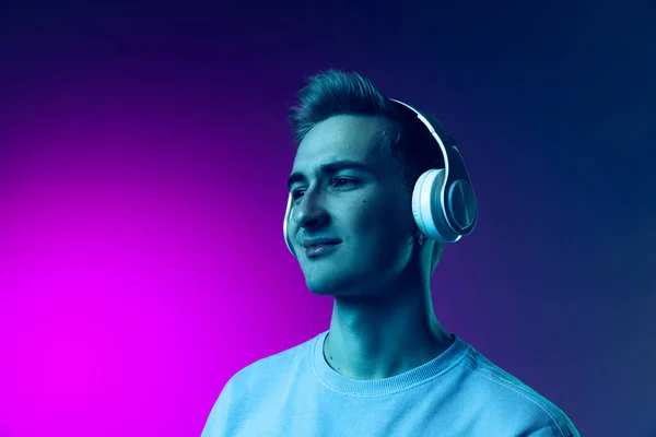 Portrait of young calm man listening to music in headphones isolated over gradient pink and purple background in neon — Stock Photo, Image