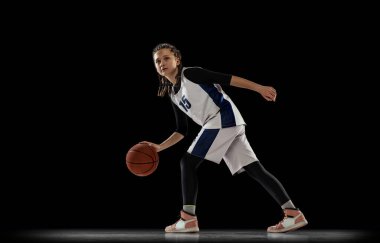 Genç bir kızın portresi, hareket halindeki basketbol oyuncusu, siyah arka planda izole edilmiş bir eğitim. Top sürme egzersizleri.