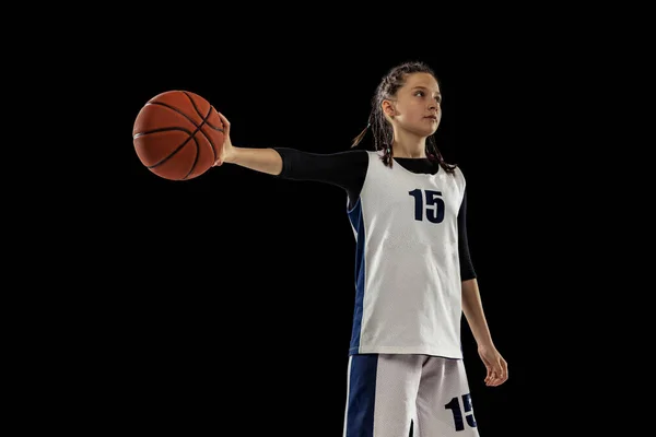 Portrait of young girl, basketball player holding ball with one hand, posing isolated over black studio background — Stockfoto