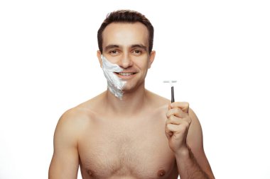 Yüzü traş köpüğüyle kaplı gülümseyen genç bir adam portresi, elinde jiletle beyaz stüdyo arka planında.