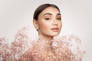 Şefkatli doğal güzellik. Çıplak makyajlı, gri arka planda küçük pembe çiçeklerle poz veren genç bir kızın portresi.